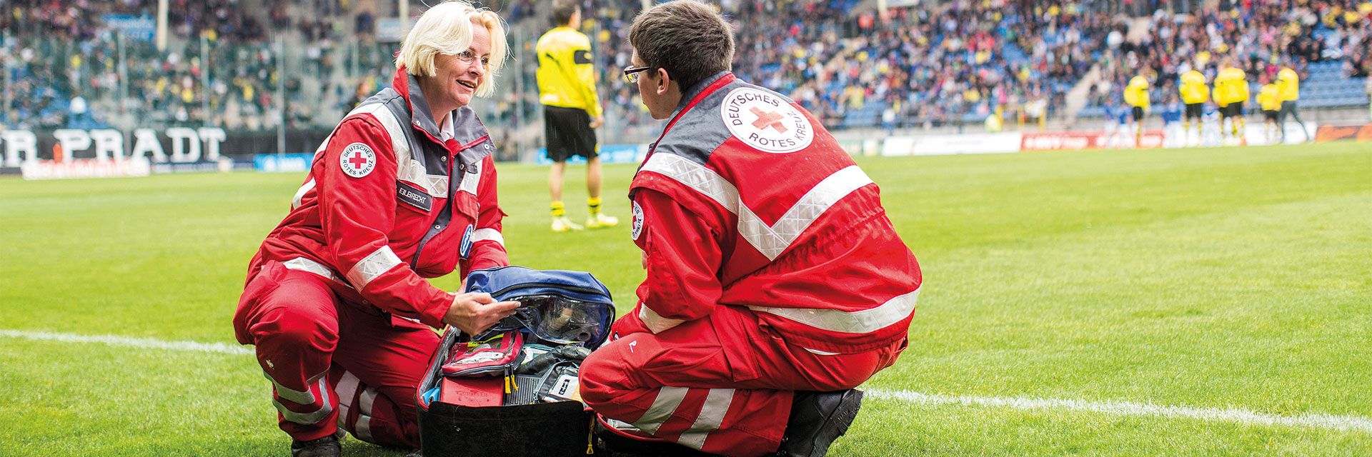 Foto: Zwei Mitarbeiter des DRK-Sanitätsdienstes tauschen sich am Spielfeldrand fachlich miteinander aus. Der Sanitätsrucksack liegt geöffnet zwischen den beiden.